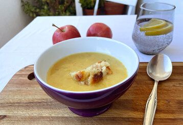 Eine Suppenschale mit Kartoffelsuppe und einem Stück Apfelkuchen steht auf einem Tisch mit einer weißen Tischdecke, daneben liegt ein Löffel und ein Wasserglas steht auch auf dem Tisch