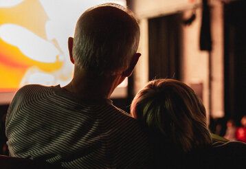 erwachsener Mann und Kind im Kino, von hinten fotografiert, Kopf des Kindes lehnt an der Schulter des Mannes, im Hintergund die leuchtende Leinwand