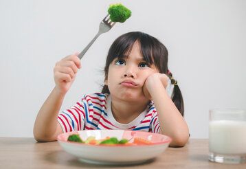 kleines Mädchen hat ein Brokkoliröschen auf der Gabel und guckt es trotzig an