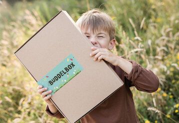 Junge steht auf einer Wiese und hält sich einen Pappkarton vors Gesicht