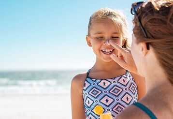 Ein Mädchen wird von seiner Mutter am Strand mit Sonnencreme eingecremt, im Hintergrund Meer