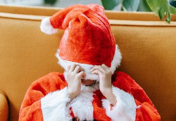 Kind mit sitzt mit einem Weihnachtsmann-Kostüm auf einem Sofa und zieht sich die Mütze übers Gesicht