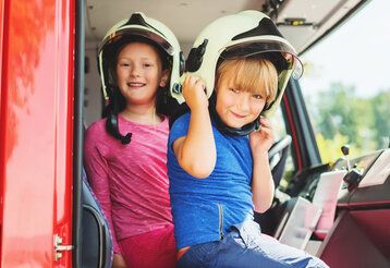 Zwei Kinder sitzen im Führerhaus eines Feuerwehrwagens, beide tragen Helme und gucken in die Kamera