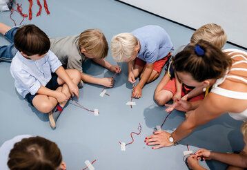 Meherere Kinder sitzen auf einem blauen Boden und basteln mit Drähten, eine erwachsene Frau hilft