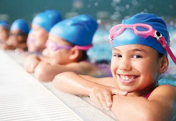 mehrere Kinder im Schwimmbecken, halten sich am Beckenrand fest, alle tragen die gleichen Badekappen und Chlorbrillen, das vordere Mädchen lacht in die Kamera