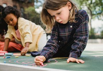 Kinder sitzen auf dem Schulhgof und malen mit Straßenkreide auf den Boden