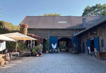 Kinderbauernhof Neuss, Blick auf Bauernhaus mit Toren, Vorplatz mit Heuballen, Sonnenschirmen, Sitzgelegenheit und Kürbissen