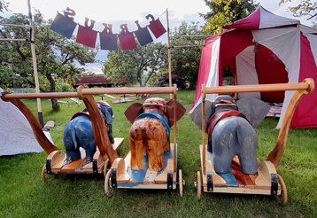 eine Wiese mit kleinen Zelten und Elefantenfiguren auf Rädern von hinten