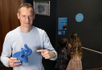 ein mann hält einen Plüschelefant in der Hand und zeigt an ihn, im Hintergrund: zwei Kinder von hinten