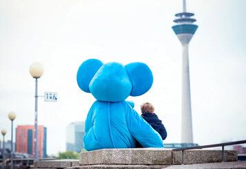 Blauer Elefant, Maskottchen des Kinderschutztbund sitzt mit kleinem Jungen vor dem Rheinturm