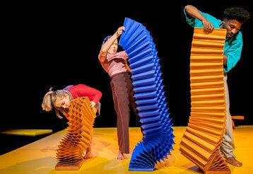 drei Schauspieler:innen spielen auf der Bühne mit überdiemensionalen, bunten, zieharmonikaähnlichen Objekten