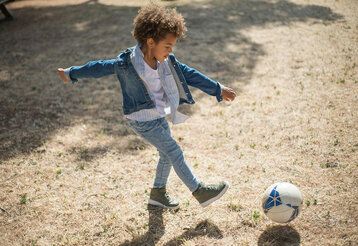 Kleines Kind mit locken und Jeans spielt auf einer trockenen Wiese Fussball