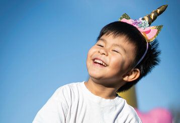 Kleiner Junge mit Einhorn-Haarreifen lächelt glücklich, im Hintergrund blauer Himmel