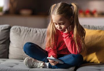 Kleines Mädchen  sitzt im Schneidersitz auf dem Sofa und daddelt am Smartphone, guckt gelangweilt