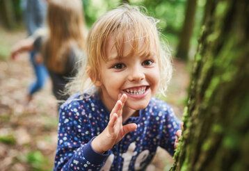 kleines Mädchen steht lachend an einem Baum im Wald