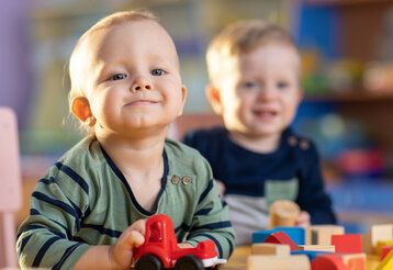 zwei Kleinkinder spielen in einer Kita mit Bauklötzen und Spielzeugauto, ein Kind ist im Vordergrund, das andere im Hintergrund unscharf