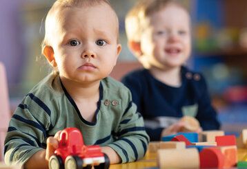 Kleinkind spielt mit Holzspielzeug und guckt skeptisch in die Kamera, im Hintergrund ein lachendes Kleinkind, unscharf
