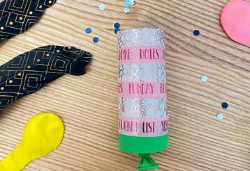 selbstgebastelte Konfettikanone auf Holztisch, daneben Federn, Luftballons und Pailletten