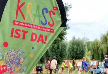 Beachflag mit Krass-Logo im Vordergrund, im Hintergrund Kinder, die an Staffeleien malen