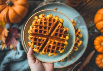 eine Hand hält einen blauen Teller, auf dem eine Waffel liegt in die Kamera, im Hintergund Kürbisse und Herbstlaub