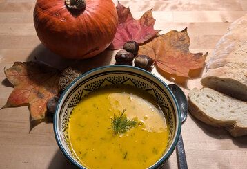 Eine Schale mit Kürbissuppe, daneben liegt ein Löffel, aufgeschnittenes Brot, Herbstlaub, Kastanien und ein Hokkaido-Kürbis
