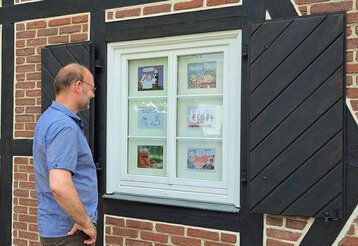 Ein Mann steht vor dem Fenster eines Fachwerkhauses, in dem Cartoons präsentiert werden, und grinst