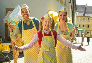 Ein Mädchen steht mit Vater und Mutter auf einem Platz, alle drei tragen Eisverkäufer-Schürzen und -hütchen und ein Eis im Hörnchen