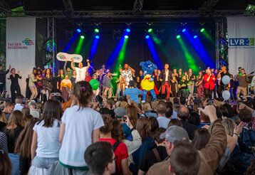 Open-Air-Bühne, auf der sehr viele Menschen singen und tanzen, avor Publikum
