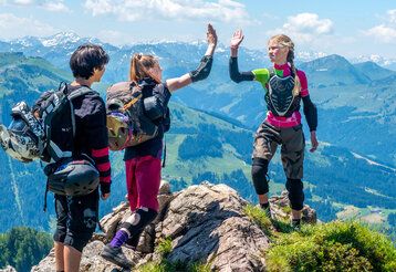 Drei Jugendliche stehen auf einem Berggipfel und machen High Five, im Hintergrund Bergpanorama