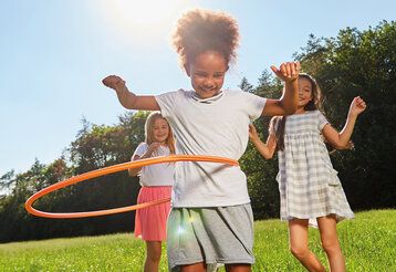 Drei Mädchen auf einer Sommerwiese, eines lässt den Hula-Hoop-Reifen kreisen