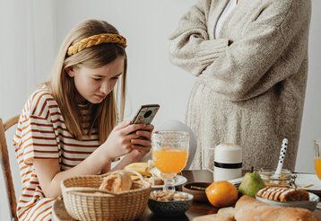 Mädchen sitzt am Frühstückstisch und daddelt, die Mutter steht mit verschränkten Armen daneben