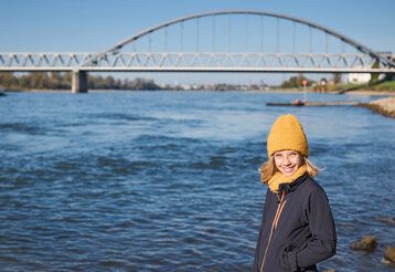 Mädchen mit Mütze steht am Rhein, im Hintergrund die Eisenbahnbrücke von Düsseldorf-Hamm