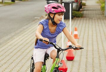Ein Mädchen mit einem pinken Fahrradhelm fährt mit dem Fahrrad einen Parcours, der mit Verkehrshütchen abgesteckt ist