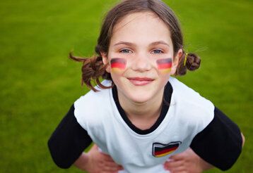 Ein Mädchen steht auf grünem Rasen und guckt in die Kamera, auf den Wangen ist die Deutschlandflagge geschminkt, sie trägt ein Deutschland-Trikot