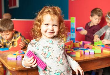 Kleines Mädchen mit einem bunten Bauklotz in Kita, im Hintergrund andere Kinder die an einem Tisch mit Bauklötzen spielen