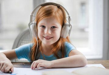 Ein Mädchen sitzt an einem Tisch und malt, dabei trägt es weiße Kopfhörerund lächelt in die Kamera