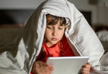 Mädchen liegt im Bett und schaut auf sein Tablet, die Betdecke liegt auf dem Kopf