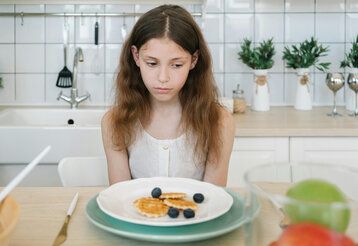 Mädchen sitzt am Küchentisch, vor sich ein Teller mit Eaffeln und Blaubeeren und  möchte nicht essen