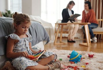 Mädchen sitzt im Wohnzimmer und spielt, zwei Frauen sitzen im Hintergrund