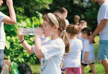 Mädchen trinkt aus Wasserflasche, im Hintergrund andere Kinder und Erwachsene ineinem Park