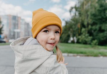 ein Mädchen mit gelber Mütze ist unterwegs auf einer Straße und dreht sich zum Betrachtenden um