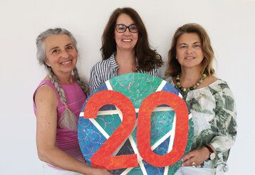 drei Frauen stehen nebeneinander und halten ein Objekt mit einer roten 20 in die Kamera