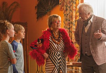 Ein Junge und ein Mädchen steh mit einer älteren Dame und einem älteren Herrn zusmmen in einem Wohnzimmer, die Dame trägt eine rote Feder-Boa, Klunker und eine rote Brille
