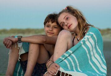 Ein Junge und ein Mädchen sitzen im Abendlicht am Strand, über den Schultern ein Handtuch