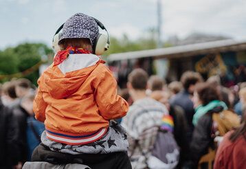 ein Kind mit Kopfhörern auf den Schultern eines Erwachsenen, von hinten, im Hintergrund Demonstrierende