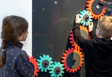 In der Ausstellung bauen Besuchende eine große Kettenreaktion. Schon bei der ersten Herausforderung muss getüftelt werden, um den Impuls vom Zahnrad an der Wand entlang weiterzugeben