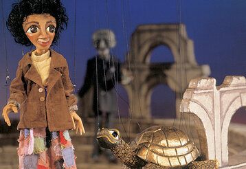 Marionette Momo steht mit einer goldenen Schildkröte auf der Bühne, im Hintergrund ein grauer Mann