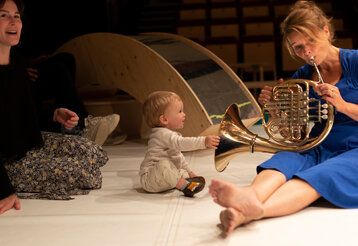 Frau im blauen Kleid spielt Horn, ein Baby guckt zu und fasst das Instrument an, beide sitzen auf der Erde, eine andere Frau sitz dabei und lacht