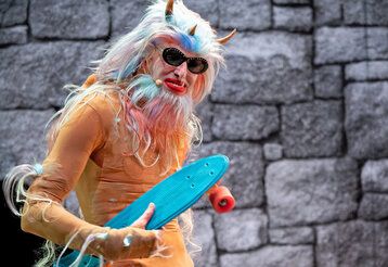 ein als Monster verkleideter Mann steht vor einer Mauer, er trägt ein kleines blaues Skateboard