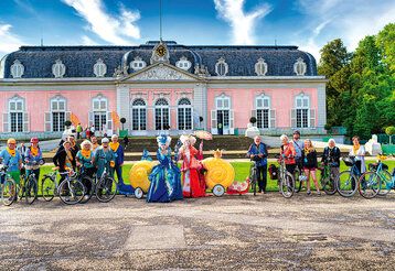Fahrradfahrer und Frauen in barocken Kostümen vor Schloss Benrath